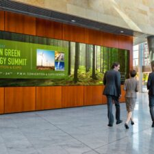 People walking by a video wall in the lobby of a building that's displaying information for a Green Energy Summit.