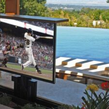An outdoor TV raised from the ground with a pool in the background.